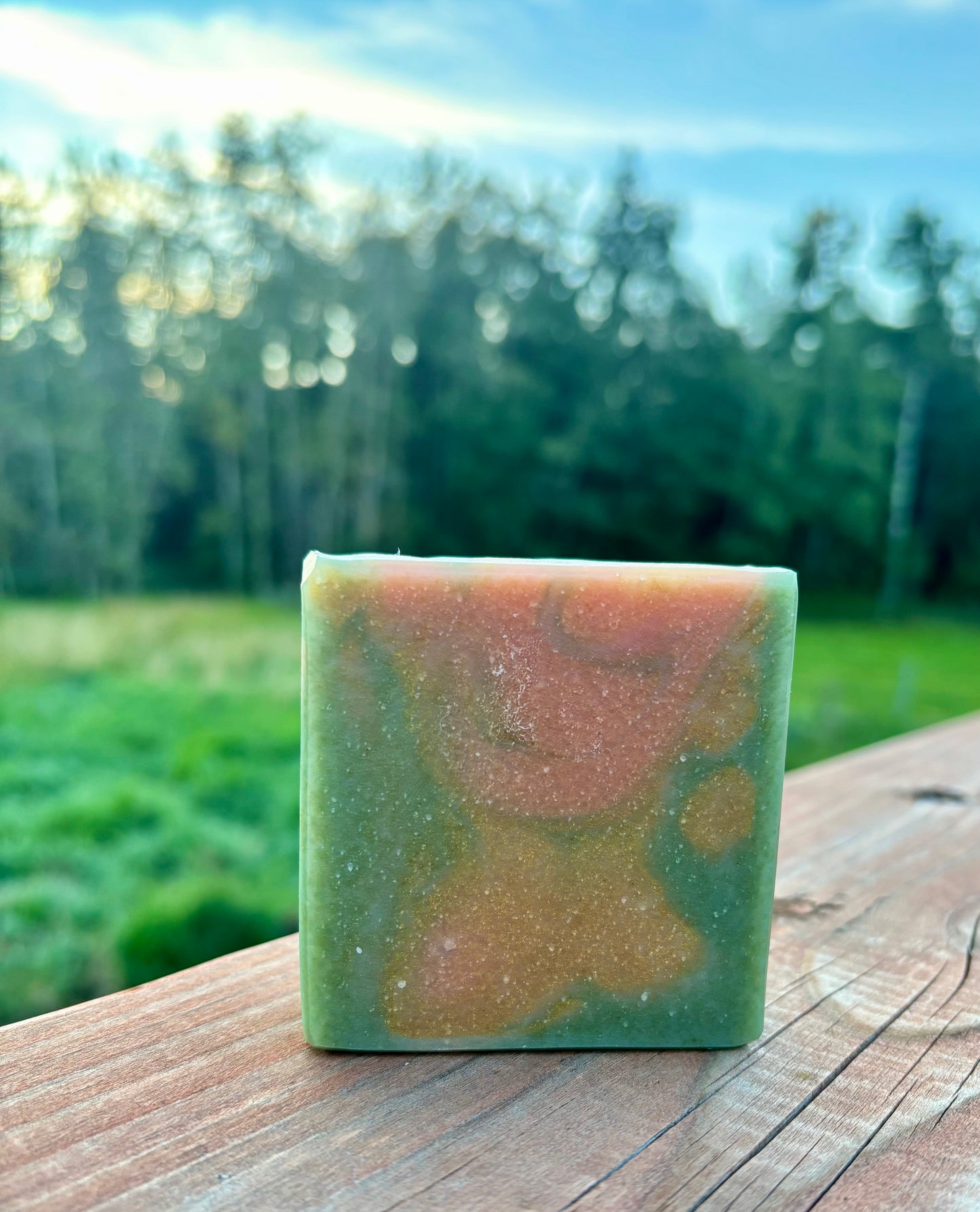 Handcrafted goat milk soap bar in earthy sage green with warm amber swirls, photographed outdoors on a wooden fence with sunlit trees — Flannel Weather by Straight Arrow Farms