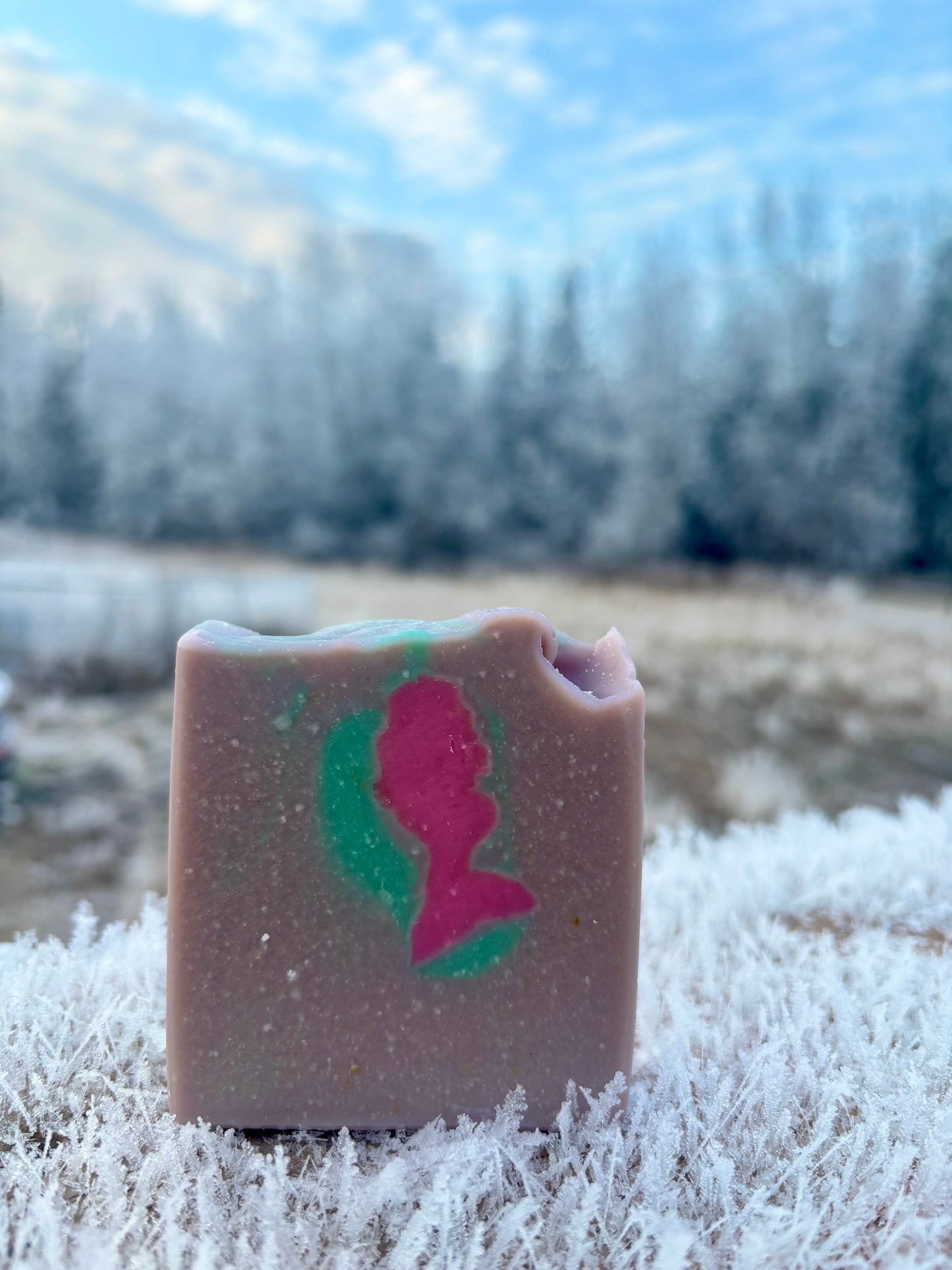 Handmade goat milk soap bar with a pink mermaid design on a sandy base, photographed on frost-covered grass with a winter landscape backdrop. Salt Water Mermaid scent by Straight Arrow Farms.
