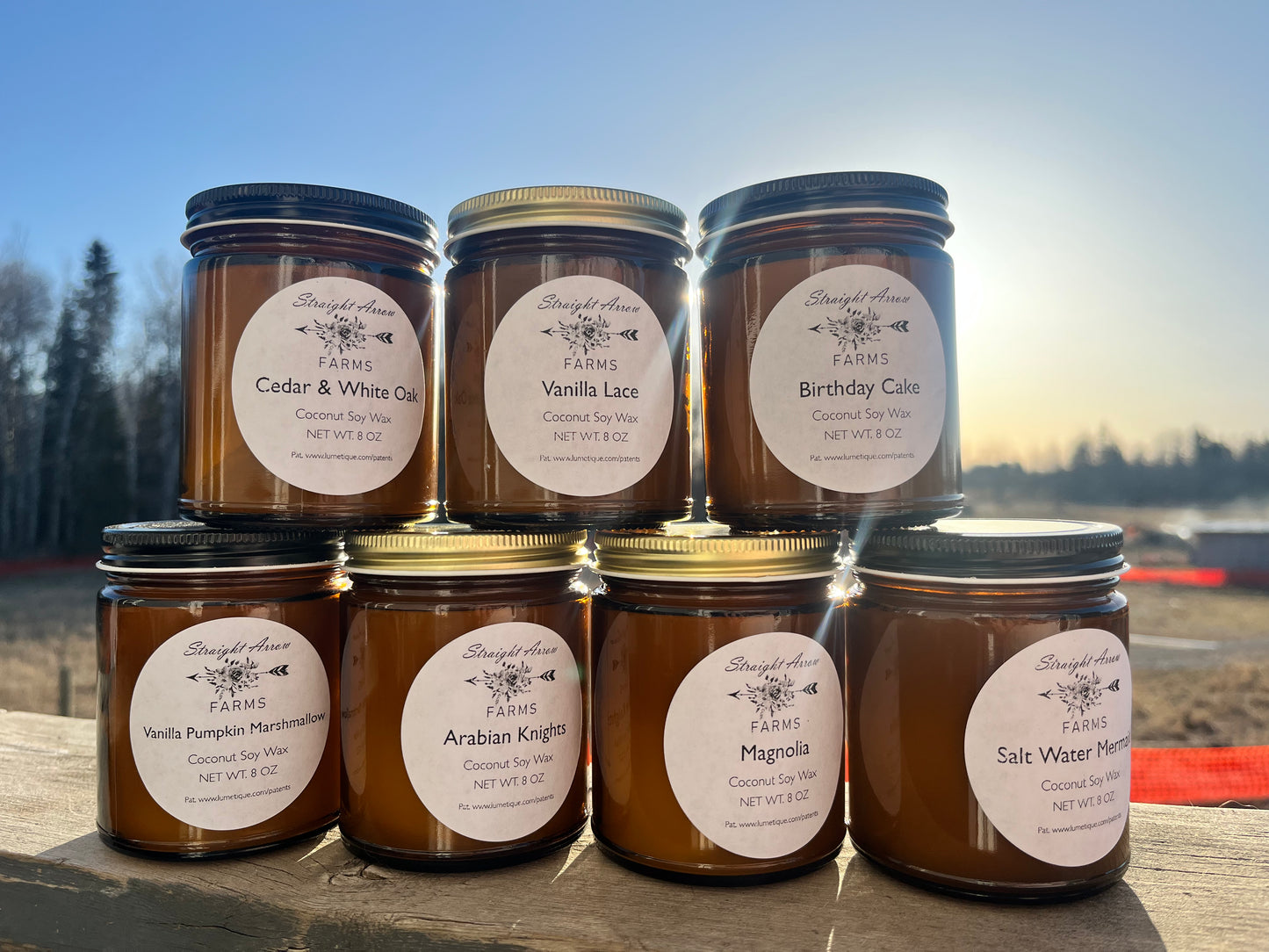 A collection of jar candles with wooden wicks, displayed in amber glass jars with various labels. They are outdoors, possibly on a wooden surface, with a bright sky in the background.