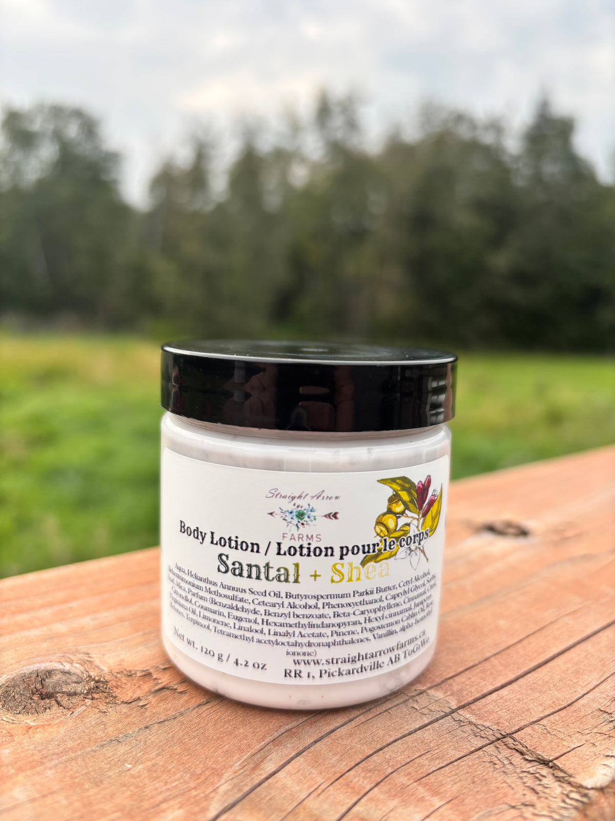 A jar of Straight Arrow Farms Santal + Shea Body Lotion sits on a rustic wooden table with a blurred outdoor forest background.