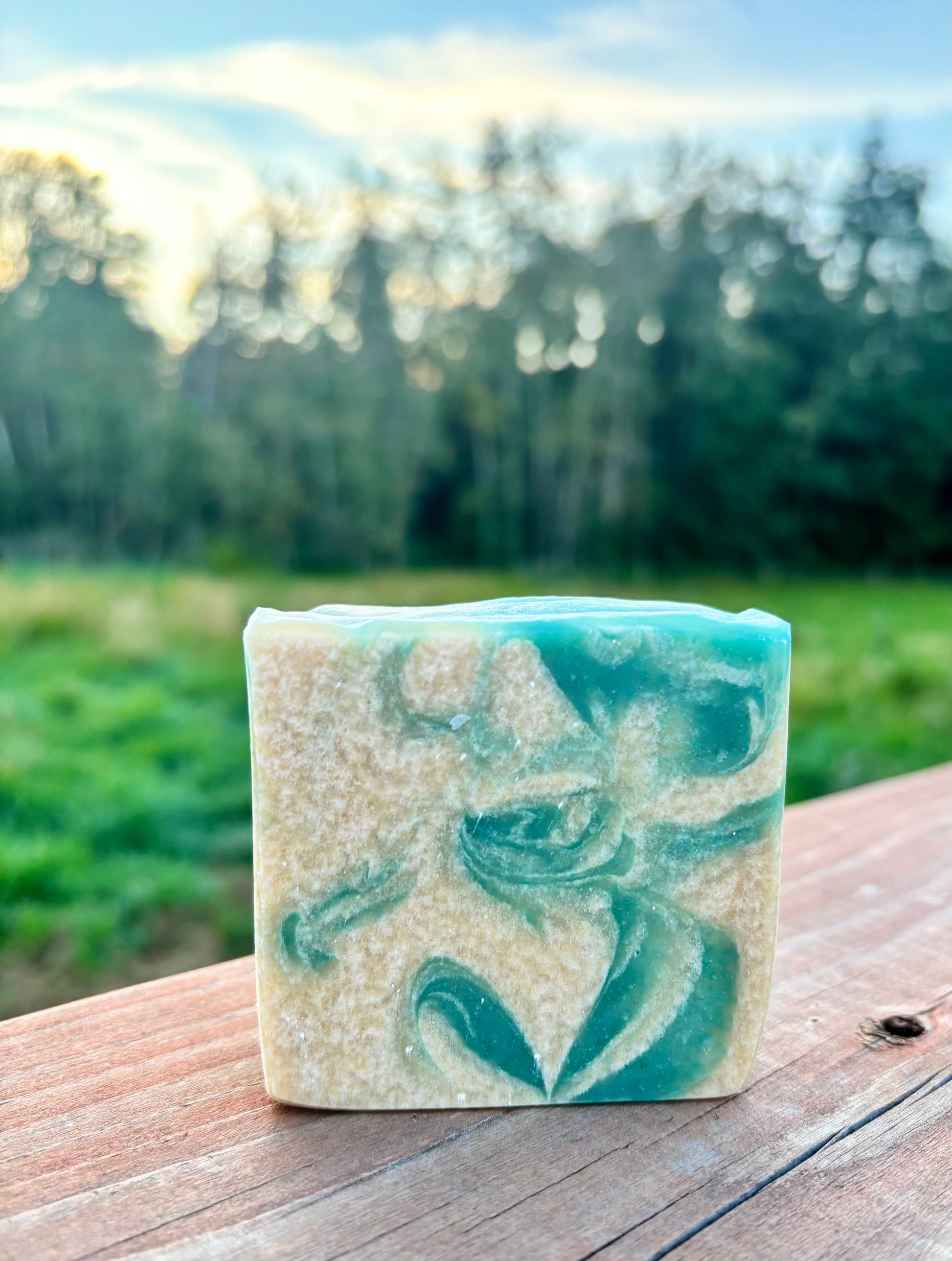 Handcrafted goat milk soap bar with teal and cream swirls, photographed outdoors on a wooden fence with a green field and trees in the background — Alpine Frost by Straight Arrow Farms
