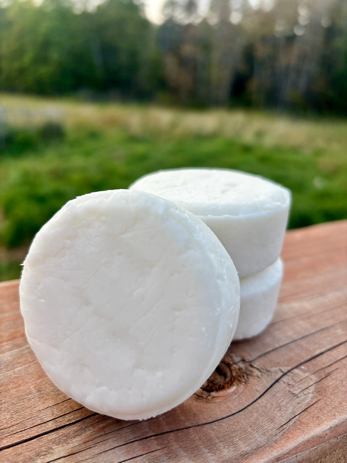 Three white plant-based shampoo bars stacked on a rustic wooden surface with a lush green farm field in the background.