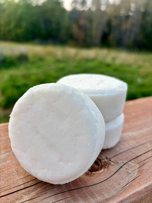Three white plant-based shampoo bars stacked on a rustic wooden surface with a lush green farm field in the background.