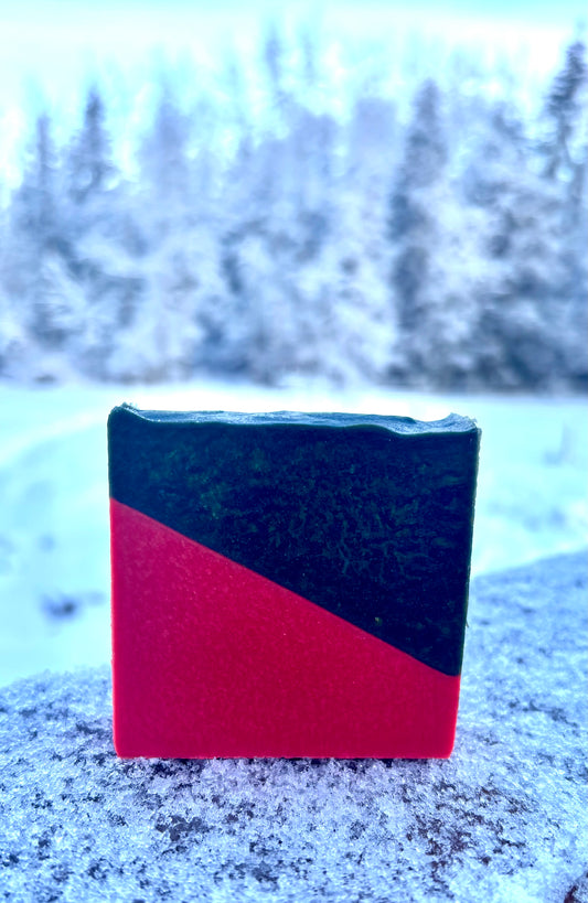Red and black rectangular goat milk soap on a snowy ground with trees in the background