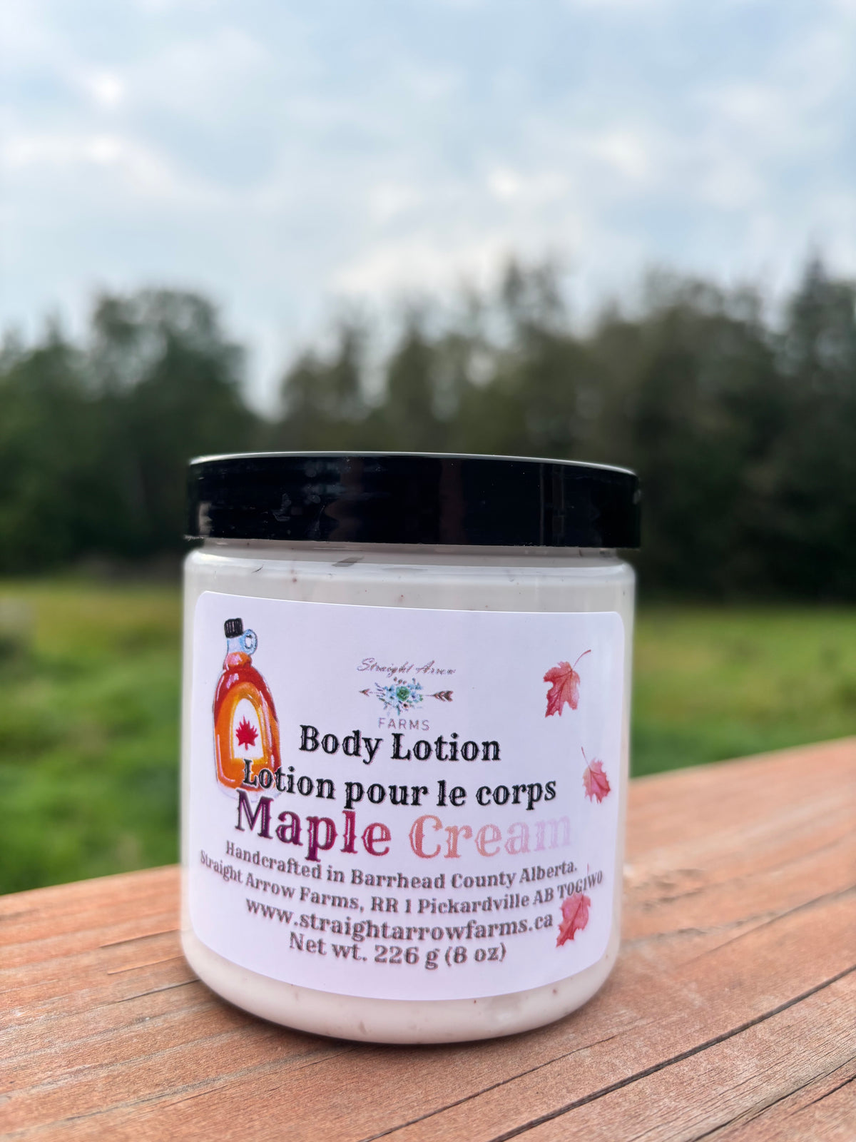 A jar of Straight Arrow Farms Maple Cream body lotion sits on a wooden table with a blurred outdoor background of green grass and trees under a cloudy sky.