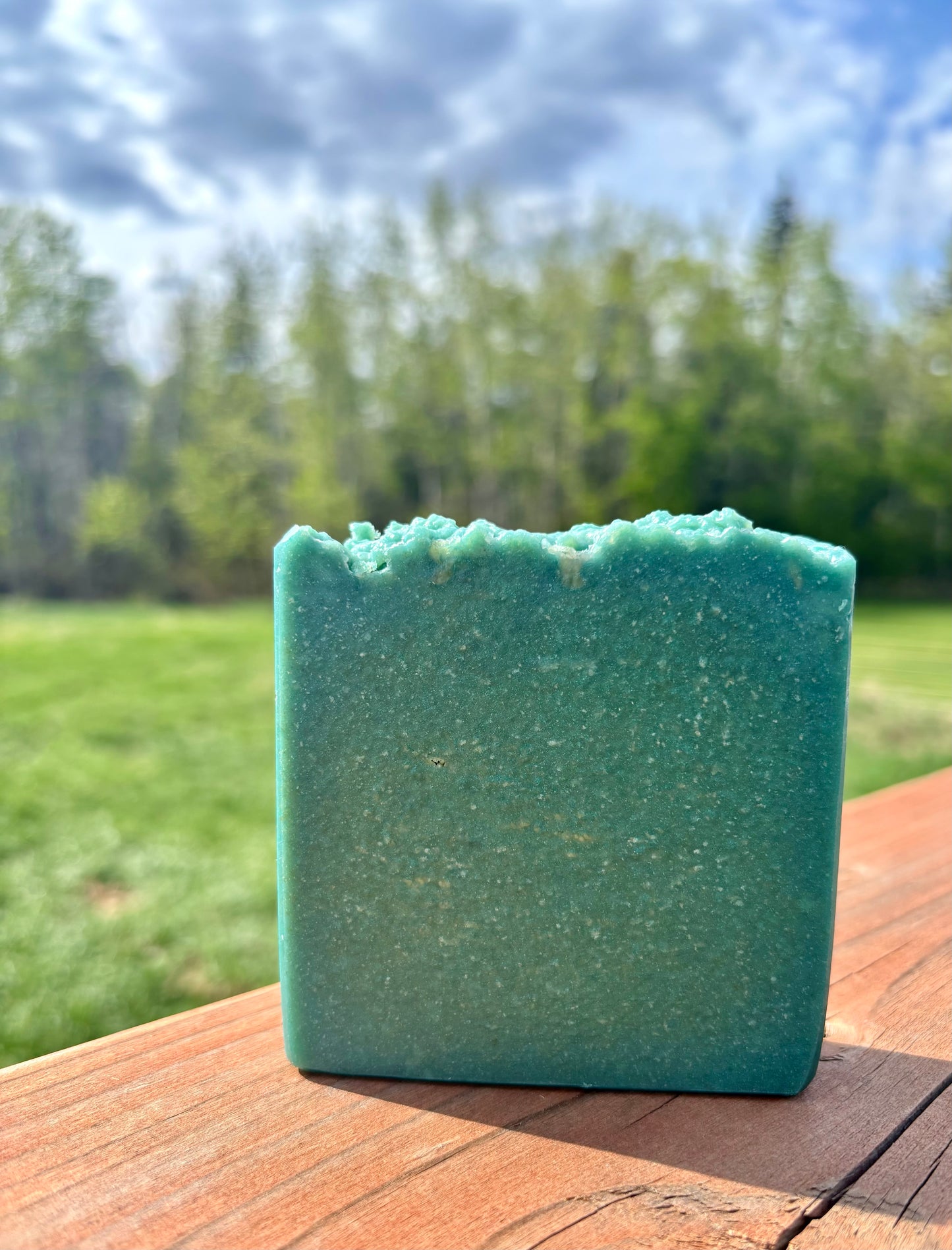 Green soap bar on a wooden surface with a natural background of trees and sky.