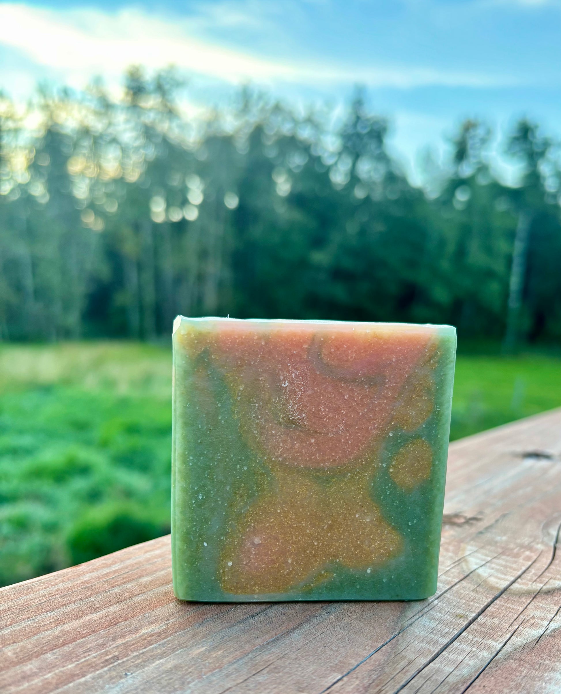 Handcrafted goat milk soap bar in earthy sage green with warm amber swirls, photographed outdoors on a wooden fence with sunlit trees — Flannel Weather by Straight Arrow Farms
