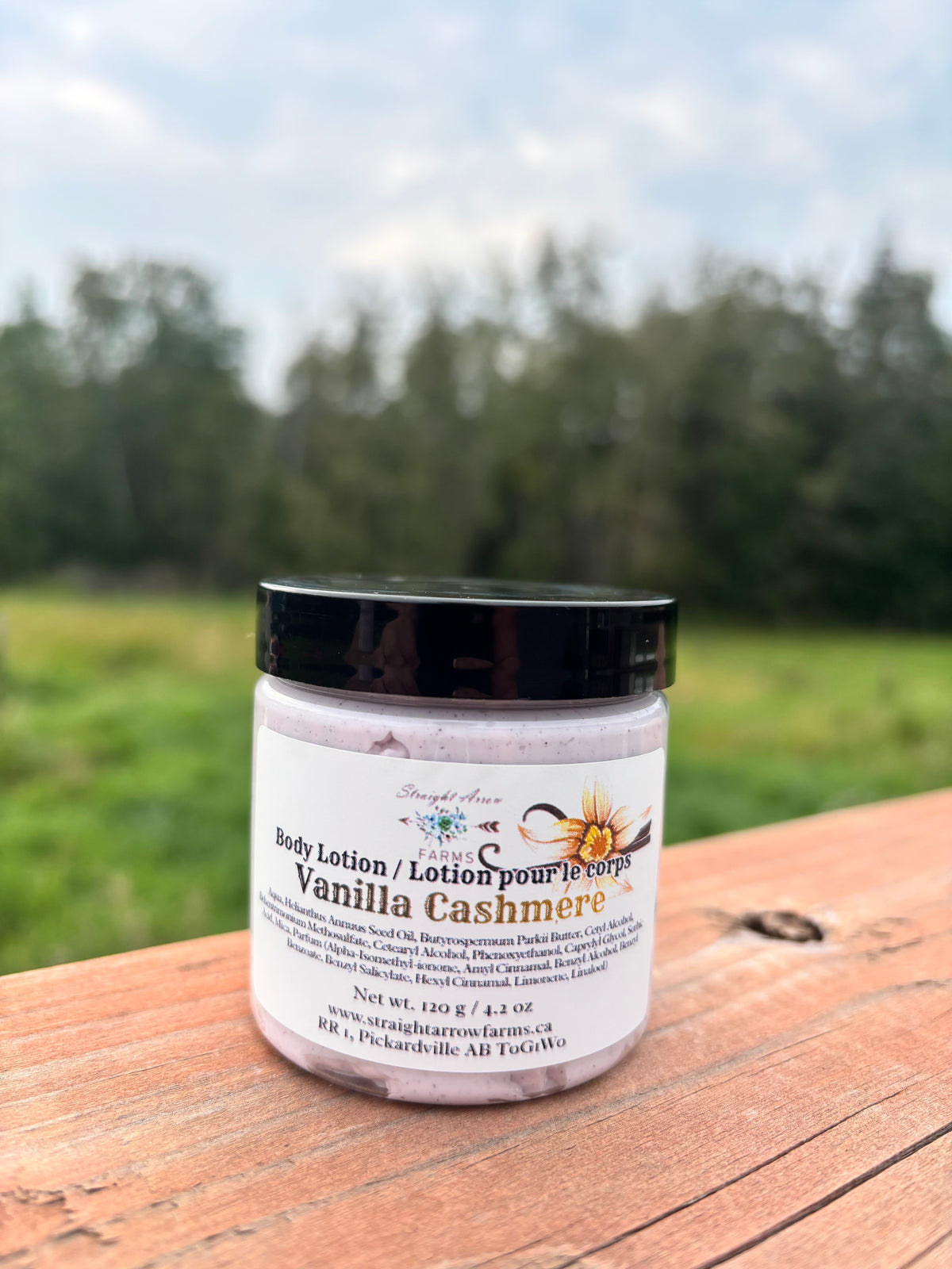 A jar of Straight Arrow Farms Vanilla Cashmere body lotion sits on a wooden deck with a blurred outdoor field and trees in the background.