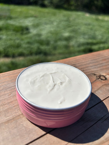 Pink tin with white grass fed tallow cream inside, on a wooden surface with a green field background