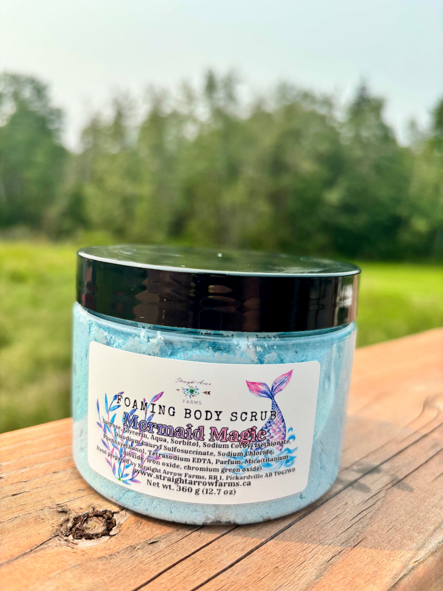 Straight Arrow Farms Foaming Body Scrub Mermaid Magic, 360g jar with black lid and mermaid tail label art, blue sugar scrub visible through jar, photographed outdoors on a wooden surface