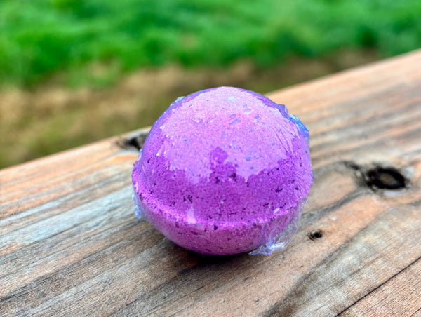 A purple bath bomb, sitting on a weathered wooden surface with a blurred green outdoor background.