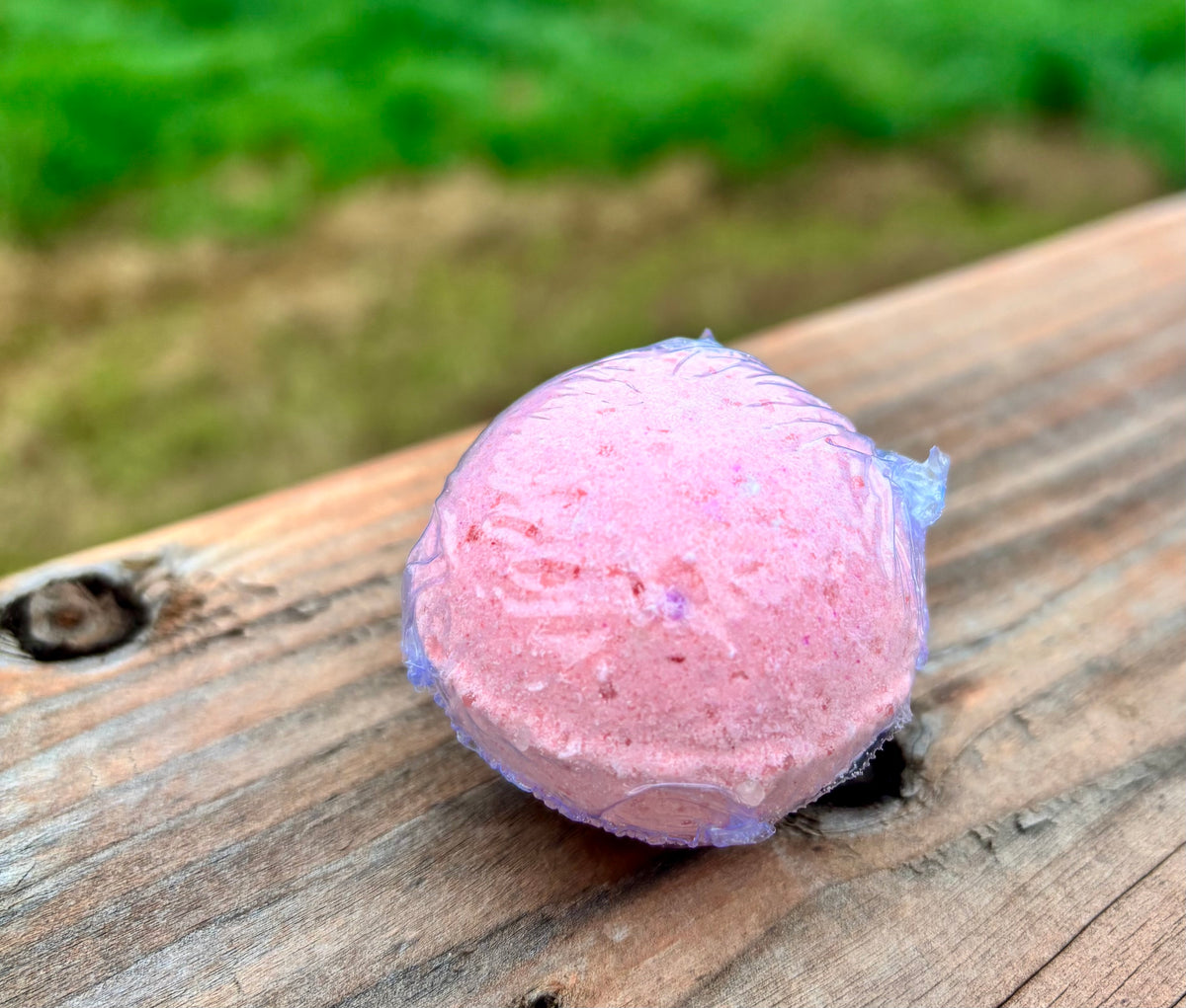 A pink bath bomb resting on a weathered wooden surface with a blurred green outdoor background.