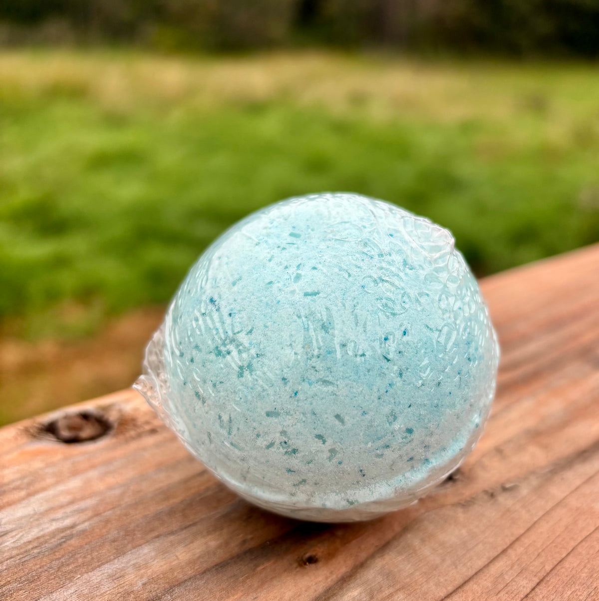 A round light blue bath bomb resting on a wooden surface with a blurred green outdoor background.
