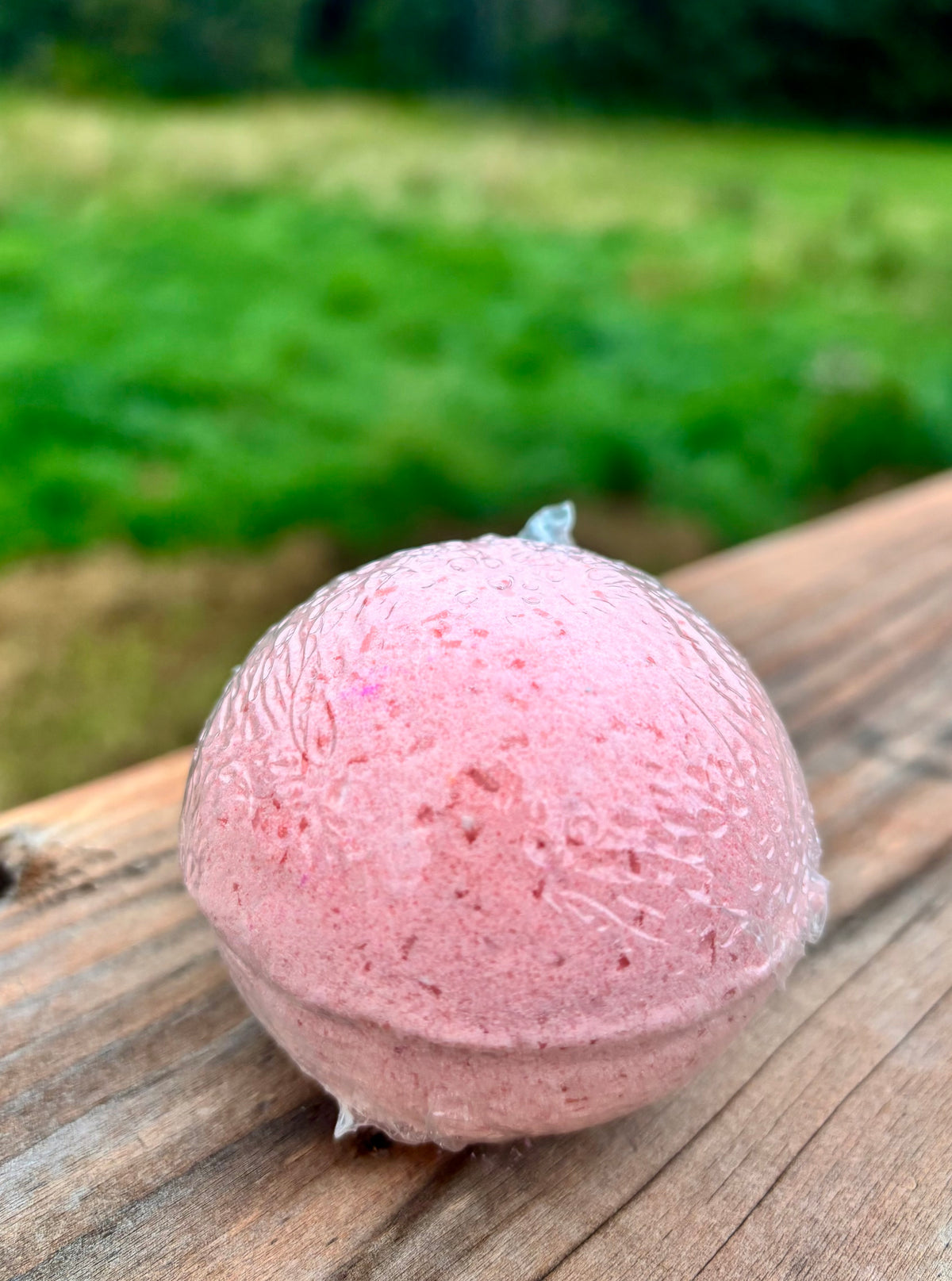 A pink spherical bath bomb resting on a weathered wooden surface with a blurred green outdoor background.