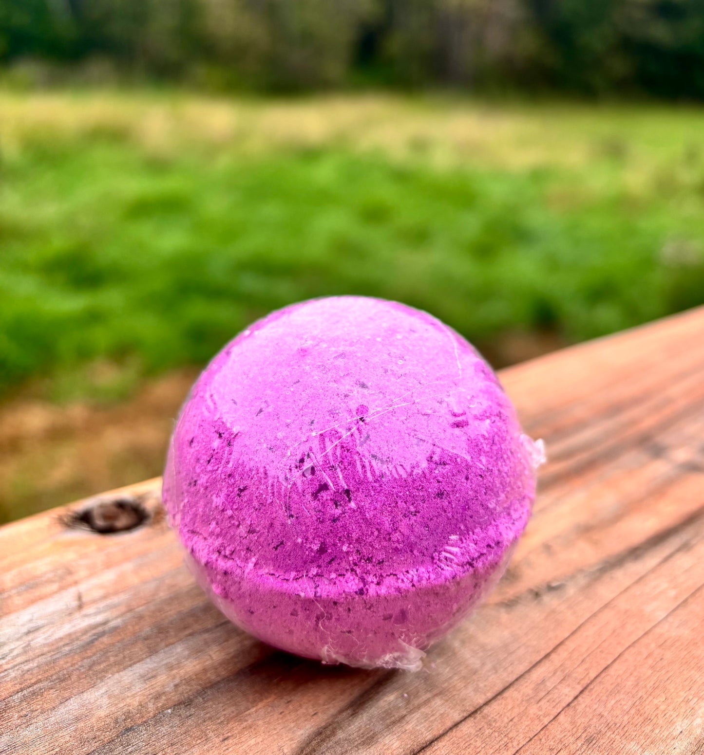 A round purple bath bomb resting on a wooden surface with a blurred green outdoor background.